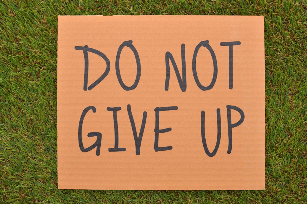 Cardboard sign reading 'Do Not Give Up' placed on green turf, symbolizing perseverance and maintaining faith.