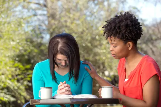Two young Christian women praying together with open Bibles, representing faith community and support while trusting God during hard times